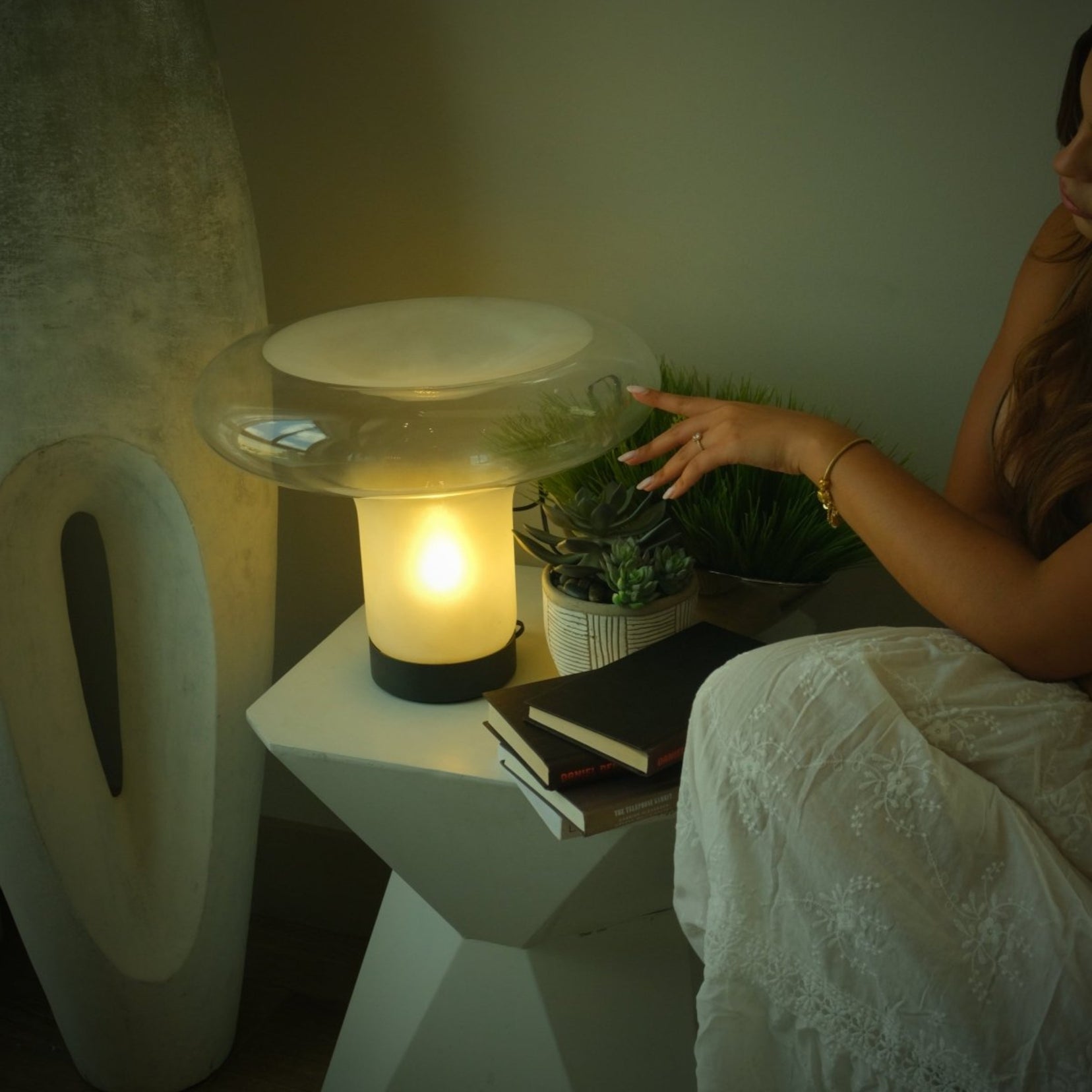 Woman sitting by a table with a Vault table lamp from Love Hohm and plant in a cozy room.